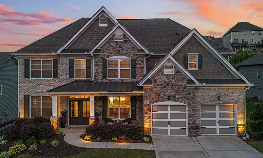 Stunning two-story brick and stone luxury home at dusk with illuminated windows, landscaped front yard, and curved walkway. Perfect real estate listing showcasing elegant architecture, inviting ambiance, and well-manicured greenery.